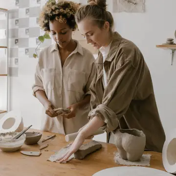 Two friends shaping clay side by side at a pottery workshop table.