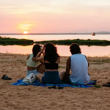 Three friends sitting on a blanket on a sandy shore, watching the sun go down over calm water.